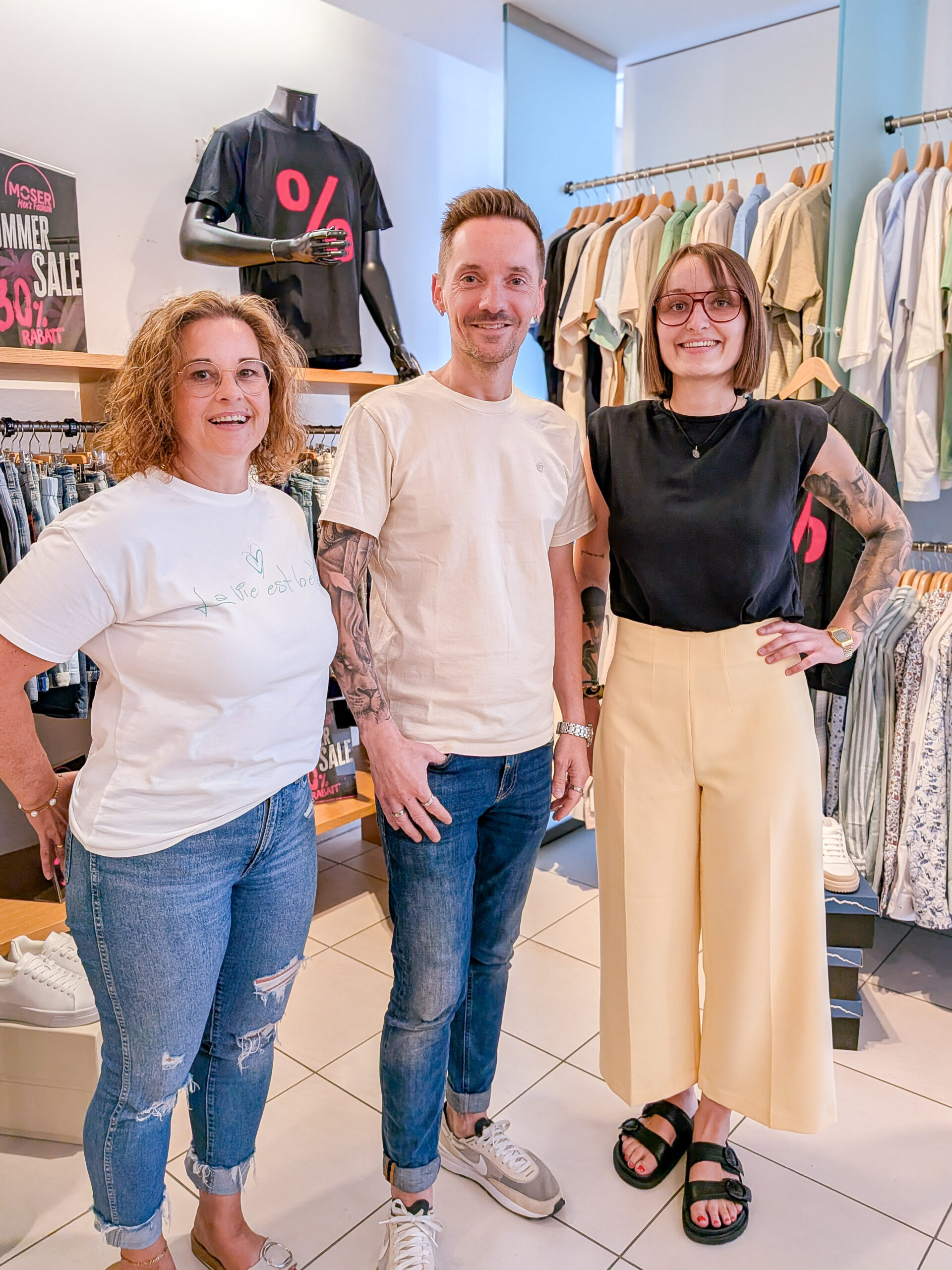 Drei lächelnde Mitarbeitende von Moser Men's Fashion vor Sommer-Sale-Kollektion im Store in Zell am Harmersbach