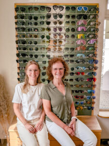 Zwei lächelnde Mitarbeiterinnen von Optik Glaser sitzen vor einer großen Sonnenbrillen-Wand mit bunter Auswahl an Sonnenbrillen in Zell am Harmersbach