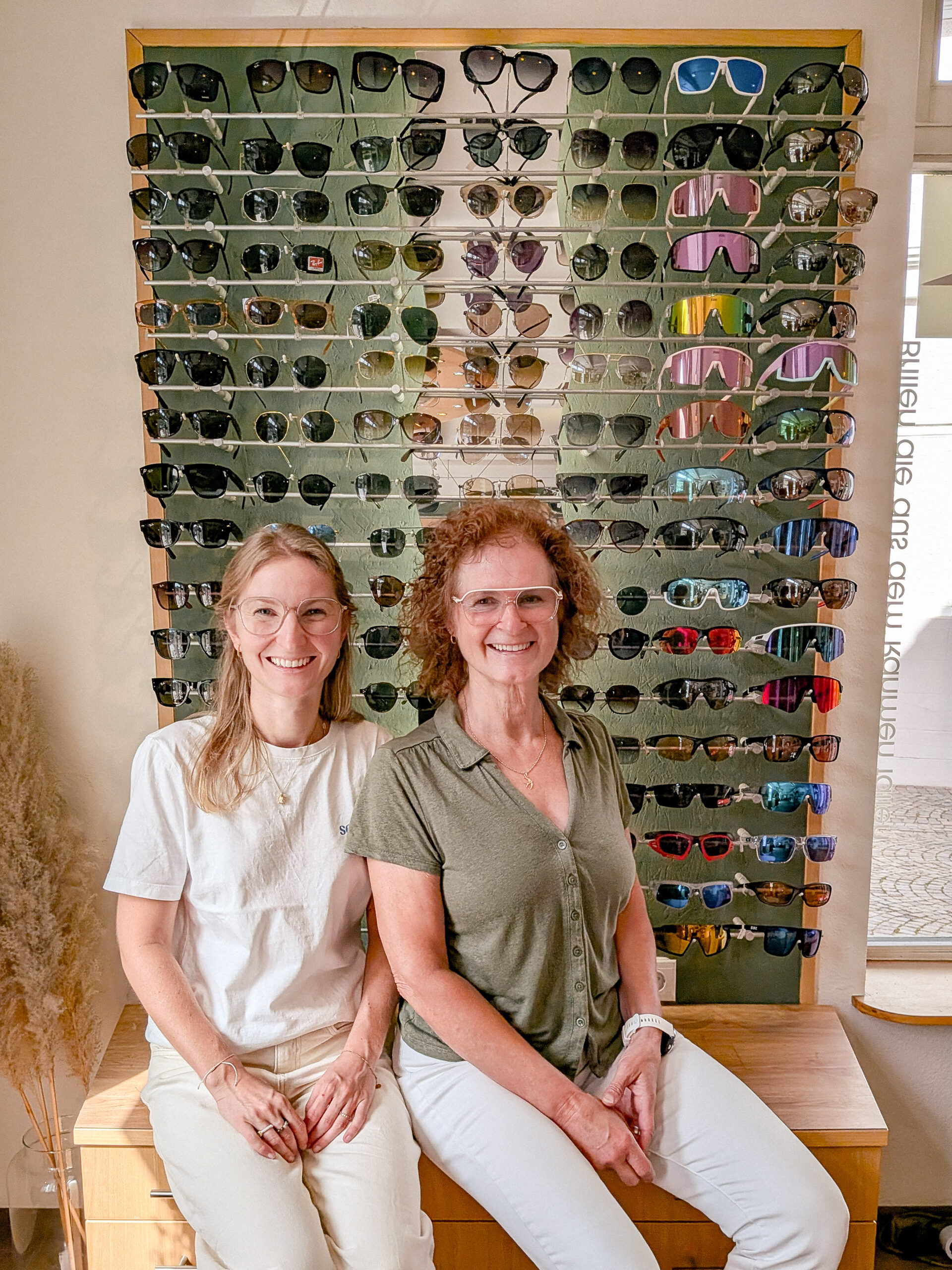 Zwei lächelnde Mitarbeiterinnen von Optik Glaser sitzen vor einer großen Sonnenbrillen-Wand mit bunter Auswahl an Sonnenbrillen in Zell am Harmersbach